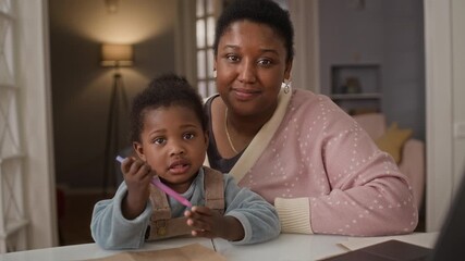Zoom in portrait of modern gen Y Black mother with two-year-old daughter sitting at table and looking at camera during educational activity