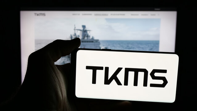 Stuttgart, Germany - 11-20-2025: Person holding mobile phone with logo of company ThyssenKrupp Marine Systems (TKMS) on screen in front of web page. Focus on phone display.