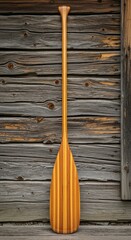 Beautiful handmade wooden canoe paddle standing vertically against a rustic weathered wooden wall outdoors for recreation