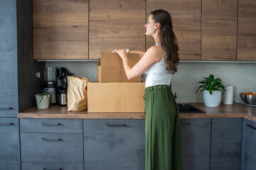 Young woman unpacking a box with pre-portioned ingredients for cooking on the kitchen counter. Concept of meal kit delivery service and easy home cooking with fresh food.