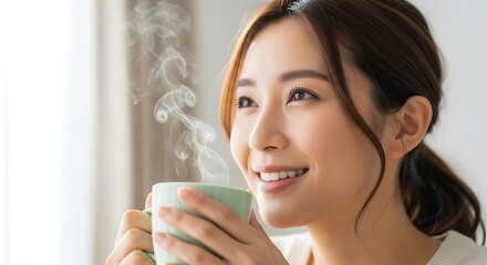 Young asian woman smiling while enjoying a warm, steaming beverage in a light green mug indoors near a window