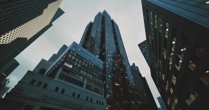 Tall buildings dominate the skyline as dusk approaches, casting reflections on the glass surfaces. The atmosphere is alive with city energy as lights begin to glow.