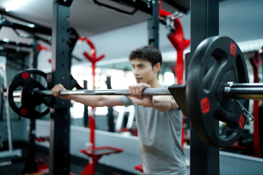young man smiles while resting chin on crossed arms over barbell, expressing peace, relief, and joy after challenging strength training session in gym