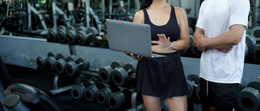 Asian man and woman exercise training in fitness gym. Young female trainer using laptop for schedule exercise training to fitness male. Workout training in fitness gym.