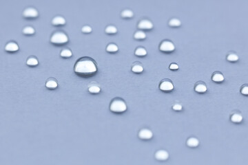 Macro close-up of clear water droplets scattered across a smooth, light blue surface. The droplets vary in size and create a soft, minimalistic pattern with shallow depth of field. 