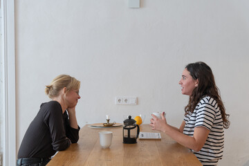 Two women sitting in the kitchen opposite each other and drinking coffee and talking