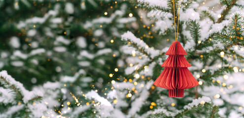 Red paper ornament hanging on snow-covered Christmas tree with golden lights. Festive composition, mockup for a New Year's card