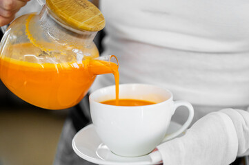 Vibrant orange juice or sea buckthorn tea is poured from a glass teapot into a white cup. Close-up on the pouring action, suggesting a healthy drink, refreshment, or warm beverage.
