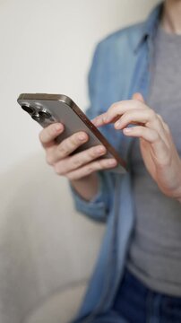 Vertical video. Close up of a female's hands holding mobile phone, scrolling her social media, messaging, browsing online content, and internet shopping indoors while sitting on a sofa at home.