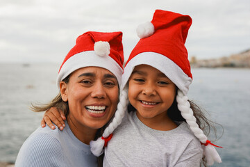Happy latin mother and daughter smiling on camera while wearing Santa Claus hats outdoor -...