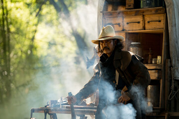 A cowboy aiming gun in a forest camp, with sunlight streaming through the smoke