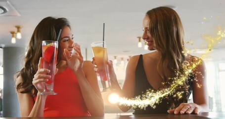 Two women at restaurant bar raising cocktails celebrating, golden spark wrapping glasses, laughing