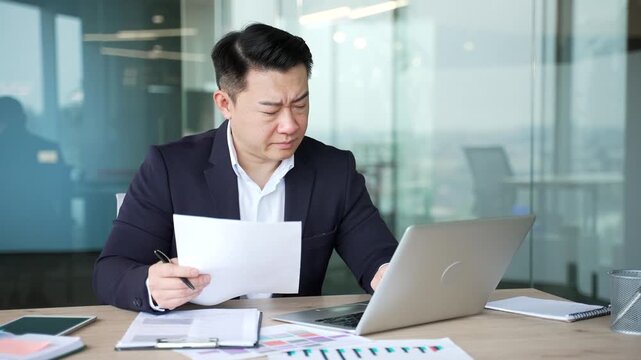Confused puzzled asian businessman having difficulty with paperwork sitting in business office. Frustrated financier unhappy with bad financial results reviewing documents and looking at laptop screen