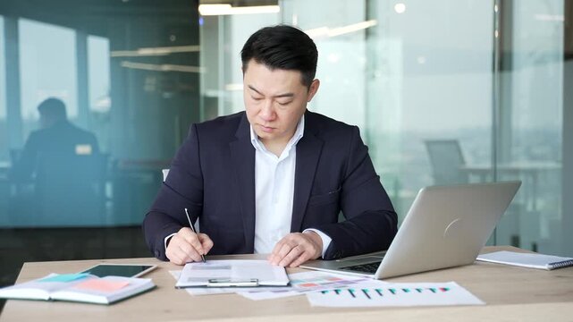 Confident asian businessman signing documents sitting at a desk at workplace in a business office. Seo in formal suit looks through the folder with documentation, checks and signs contract with a pen