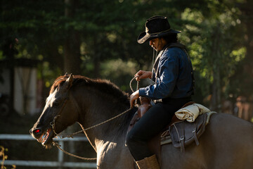 A cowboy riding a rearing horse in a fenced area surrounded by lush greenery