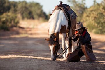 A cowboy kneeling on the ground while aiming a rifle beside his horse in a rugged outdoor setting