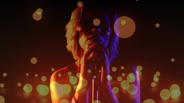 Teen singer lowering hands, revealing face and bringing mic closer to sing, rim-lit bokeh drifting