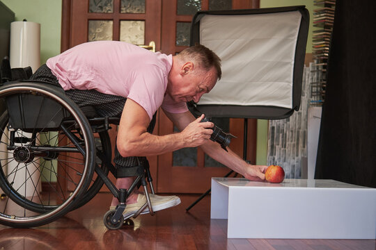 A man in a wheelchair holds a camera in his right hand and positions an apple with his left hand to take a photo of it. - Powered by Adobe