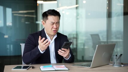 Shocked worried asian businessman reads bad news on mobile phone while sitting at desk at workplace in business office. Frustrated stressed man in formal suit received a negative message on smartphone