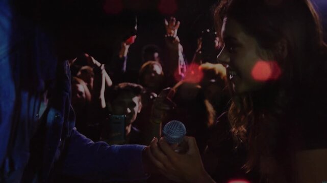 Male host offering mic, female singer grabbing mic and preparing to perform amid club bokeh