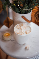 Cozy white mug filled with creamy beverage topped with marshmallows, placed on a marble table beside a lit candle and decorative greenery, creating a warm atmosphere