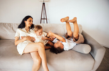 Mother with dark hair sits on a light sofa, joyfully interacting with her two children, one toddler and one young boy, creating a warm family atmosphere in a cozy living room