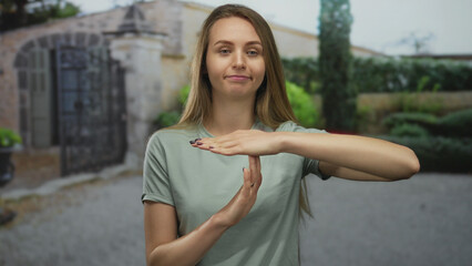 Woman making timeout gesture outdoors in garden setting with relaxed expression and light clothing,...