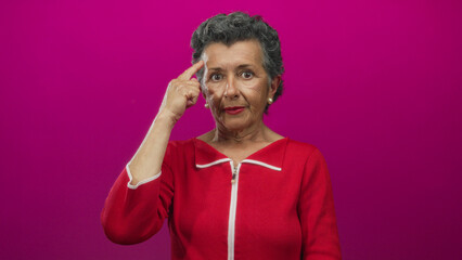 Fototapeta premium Senior woman with grey hair in red top gestures thoughtfully, pointing to her head against a vibrant pink background