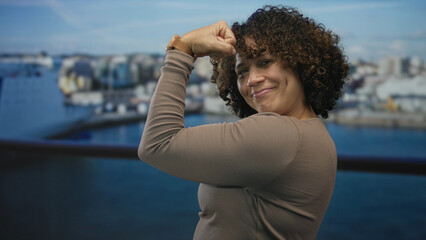 Hispanic woman flexes bare bicep aboard a boat deck at port in daylight, smiling with closed eyes; confidence.