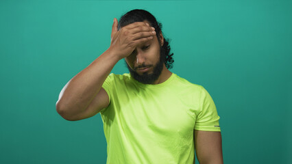 Man pinching bridge of nose in studio with teal backdrop and neon green tshirt, hand covering eyes in weary pose; stress fatigue recovery.