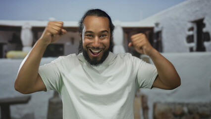 Naklejka premium Man raises fists in celebration at a building entrance, smiling widely with bare forearms and white tshirt, ponytail and beard visible; victory joy.