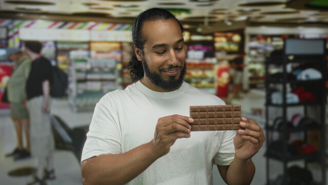 Man holds chocolate bar with both hands in busy airport terminal while examining the snack and gently smiling; curiosity. - Powered by Adobe