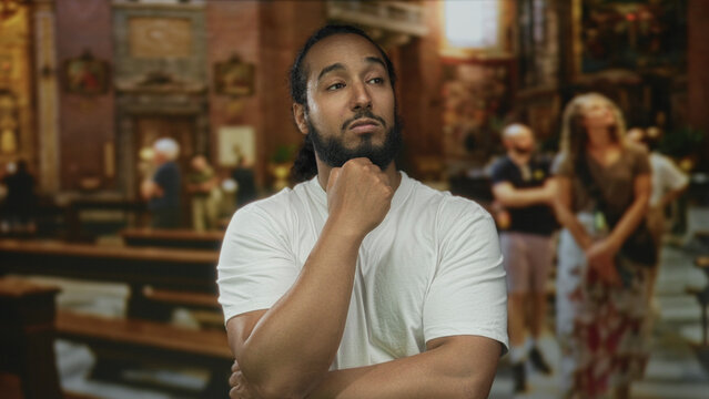 Man with hand on chin in church building, wearing white t shirt and beard amid pews and visitors; contemplation reflection.