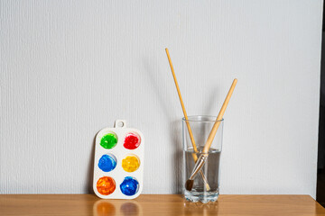 Paintbrushes in a jar of water with a palette of watercolour paints against a wall, minimal clean artist or hobby image