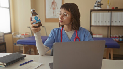 Woman in blue scrubs holds medicine bottle while examining label at clinic desk with laptop and stethoscope; care.