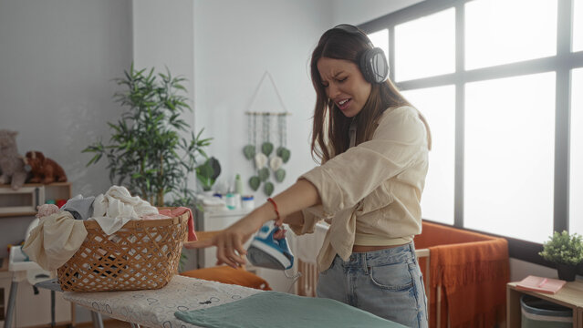 Woman ironing with steam iron on ironing board next to laundry basket and pointing finger near crib in baby room while wearing headphones and grimacing; frustration and multitasking.