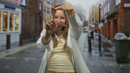 Fototapeta premium Young hispanic woman framing a square with her hands and smiling on a wet city street, midriff exposed; creative joy.