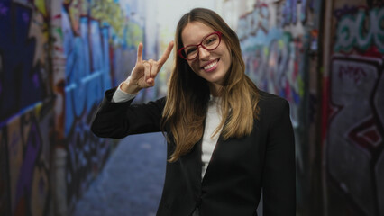 Young woman smiling and making rock horns with her hand while wearing red glasses in a graffiti street alley; playful confidence.