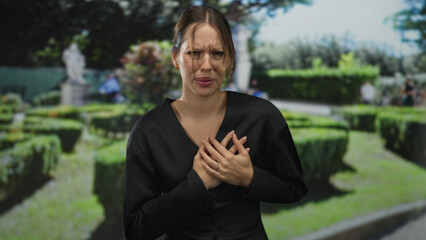 Fototapeta premium Woman clutching chest for heartattack in forest pathway beside hedge with hands pressed to sternum and grimace; medical emergency pain.