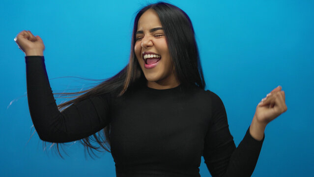 Woman celebrating joyfully in front of a vibrant blue background, embodying youthful exuberance and latin heritage against an isolated, colorful wall. - Powered by Adobe