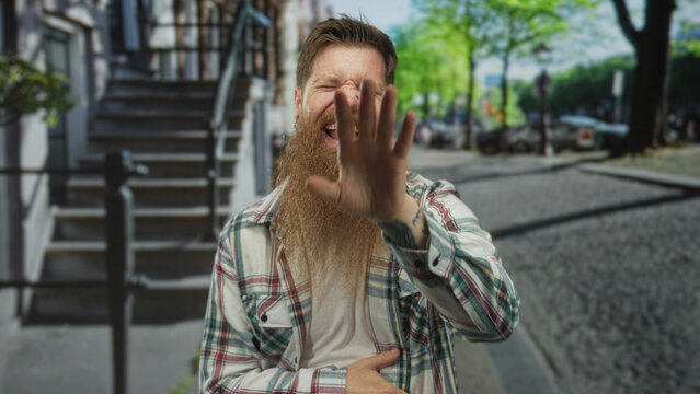 Man with long beard and plaid shirt pointing finger toward camera in urban street with building facade visible; amusement.