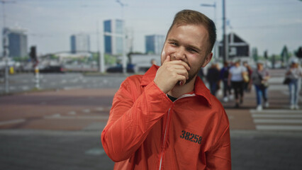 Young caucasian man prisoner hand covering mouth laughing on city street, orange jumpsuit with...
