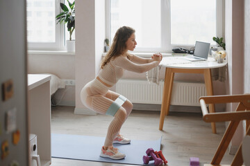 Woman exercising at home, performing squats on yoga mat with resistance band, cozy apartment interior and fitness equipment