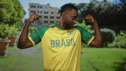 Man in yellow and green brasil jersey flexes biceps in a sunlit park with a building and potted...