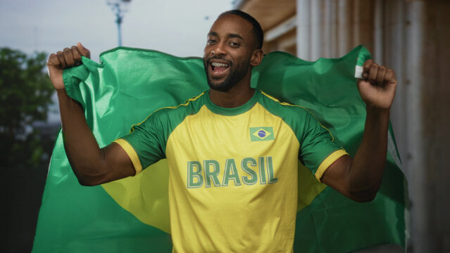 Young man wrapped in brazil flag smiling with arms raised holding the flag and wearing a brasil jersey on a city street; joy pride support.