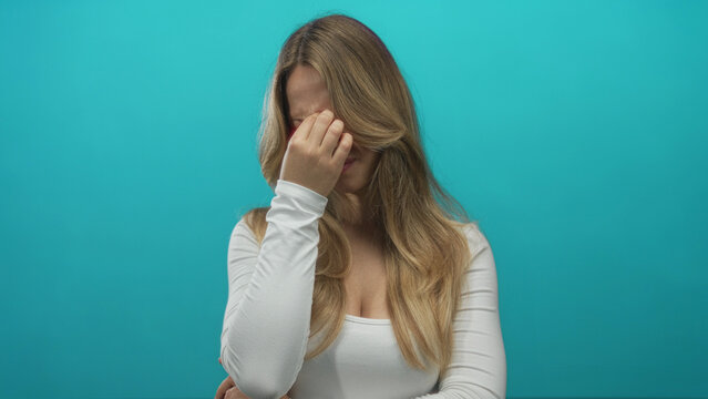 Young caucasian blonde woman pinches nose bridge under studio lights; stress headache fatigue tension.