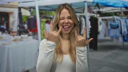 Woman blonde sticks tongue out flashing rock horns with both hands at a street market; fun playfulness rebellion.