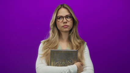 Blonde woman wearing glasses holds ornate book against chest with arms in studio; contemplation focus.