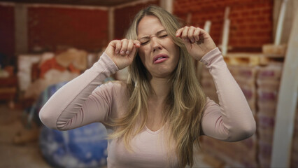 Woman with blonde hair rubs eyes against brick wall in construction site interior; sadness fatigue...