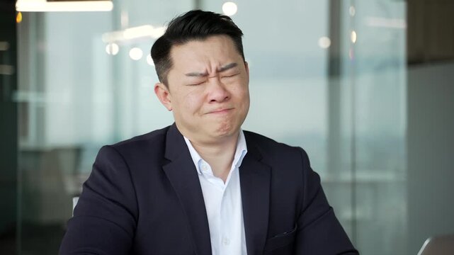 Asian businessman suffering from headache while sitting at workplace in modern business office. Unhealthy exhausted tired worker in formal suit holding massaging his temples with his hands. Close up